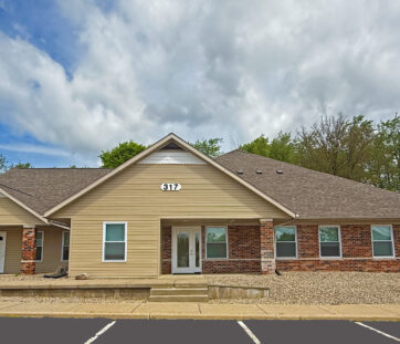 Single-story building with tan siding and brick, white trim, and glass doors. There’s a small porch out front and a mostly empty parking lot.