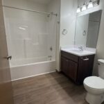 Simple bathroom with a bathtub-shower combo, wooden floor, white toilet, vanity with sink, mirror, and bright lights above the sink.