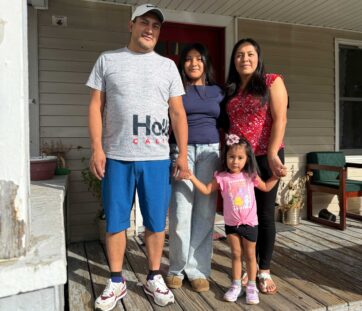 A family of four stands smiling on a porch, enjoying a sunny day together.