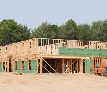 A wooden house frame is being built on a clear day, with trees in the background and some construction equipment nearby.