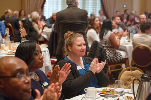 People sitting at round tables, clapping and enjoying a meal together during a lively indoor event or conference.