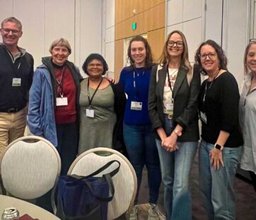 A group of six people smiling and posing together indoors at what looks like a conference or event.