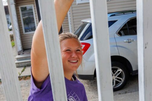 Smiling person in a purple shirt painting a white fence outside near a house and parked car.