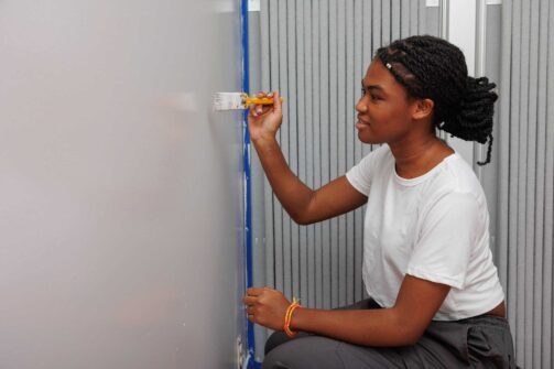 A woman in a white t-shirt paints the edge of a wall with a brush, using painter’s tape for clean lines.