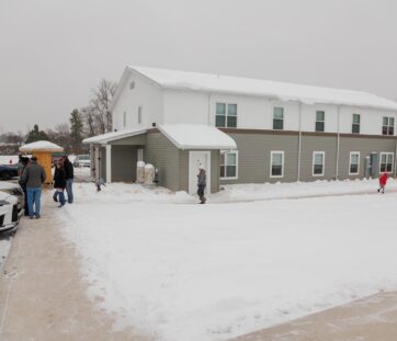 A group of people stand outside a two-story building on a snowy day, with snow covering the ground and roof.