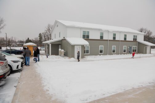 A group of people stand outside a two-story building on a snowy day, with snow covering the ground and roof.