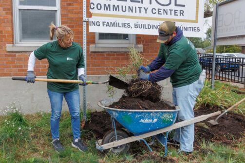 Two people in green shirts are shoveling dirt into a blue wheelbarrow outside a community center.