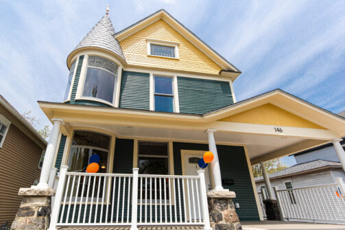 A charming two-story house with yellow and green siding, a cool turret, front porch, and some orange and blue balloons hanging by the entrance.