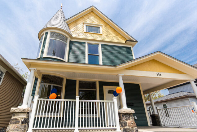 A charming two-story house with yellow and green siding, a cool turret, front porch, and some orange and blue balloons hanging by the entrance.