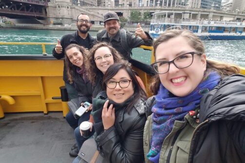 A group of friends smiling and posing for a selfie on a boat ride, with city buildings and water in the background.