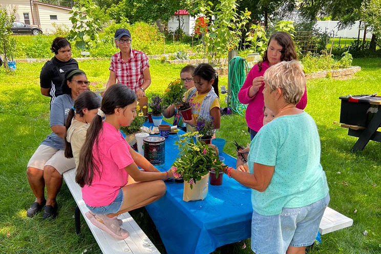 A group of kids and adults hang out at a picnic table outside, planting flowers together on a sunny day.