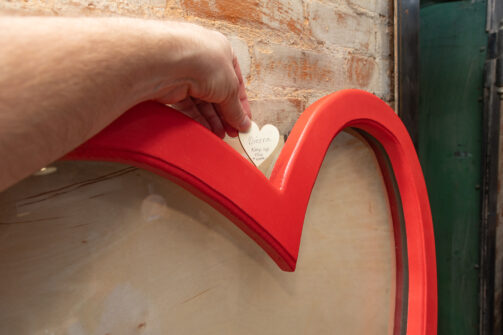 A hand places a small heart-shaped note inside a large red heart frame against a brick wall.