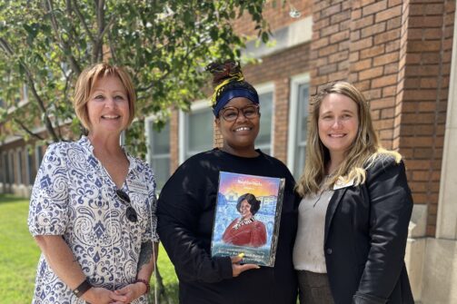 Three people stand outside smiling; the person in the middle holds up a colorful portrait artwork.