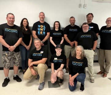 A group of people wearing matching black t-shirts posing together in a bright room, smiling for the camera.