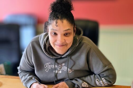 A woman in a hoodie sits at a desk, smiling and filling out paperwork in a cozy, warmly colored office space.