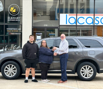 Three people standing in front of a gray SUV outside a dealership, smiling and shaking hands, looks like a car purchase or delivery.