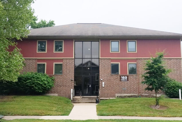 A two-story brick and red building with a glass entrance, surrounded by green grass, trees, and bushes, located at 169 State St.
