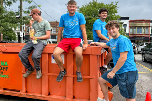 Four young guys hanging out, some sitting on an orange dumpster, all looking relaxed and happy on a cloudy day in a neighborhood.