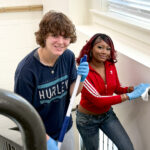 Two people smiling while cleaning a stairwell, wearing gloves and casual clothes, enjoying the task together.