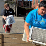 Two people are carrying old air conditioner units outside on a sunny day, getting ready to haul them away.