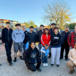Group of people posing outdoors, dressed casually with some in hoodies and jackets, smiling for the camera on a sunny day.