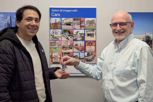 Two men smile as one hands over a key, standing in front of a poster with car images and a blue "Select all images with Cars" heading.