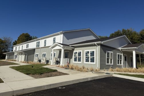 A modern, two-story apartment building with gray siding sits under a clear blue sky, surrounded by fresh landscaping and a new sidewalk.