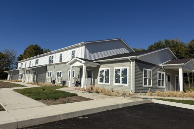 A modern, two-story apartment building with gray siding sits under a clear blue sky, surrounded by fresh landscaping and a new sidewalk.