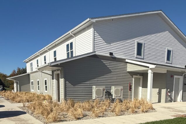 Modern gray and white apartment building with tall dry grass landscaping, under a clear blue sky on a sunny day.