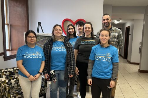 A group of six people, most wearing matching blue shirts, stand together indoors and smile for the camera.