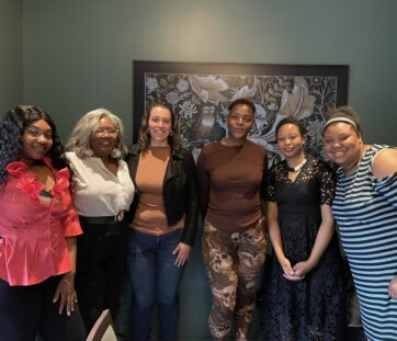Six women standing together indoors, smiling at the camera with a colorful wall art behind them.