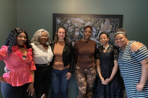 Six women standing together indoors, smiling at the camera with a colorful wall art behind them.