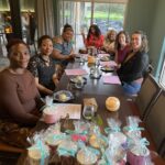 A group of women sitting around a table with snacks and gift bags, smiling and enjoying a casual get-together inside a cozy room.