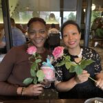 Two women sitting at a table in a restaurant, smiling and holding pink roses.
