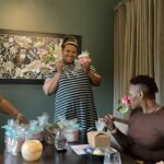 Three people hanging out at a table, smiling and holding flowers and gift bags, with a painting and curtains in the background.