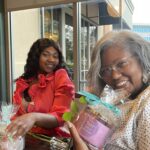 Two women sitting at a table, smiling and holding gift baskets, enjoying a casual moment together indoors.