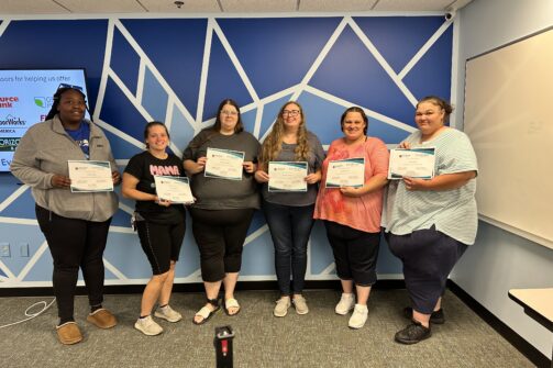 Six women standing in a row, smiling and holding certificates, in front of a blue geometric wall.