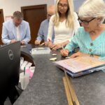 Four adults standing at a counter, filling out forms and writing on papers, with office supplies and a laptop visible nearby.