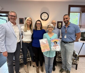 A group of six adults smiling indoors, with one woman in front holding up colorful drawings.