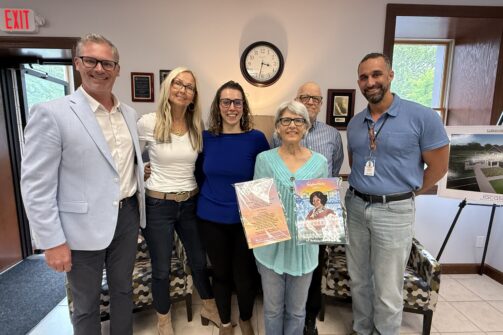 A group of six adults smiling indoors, with one woman in front holding up colorful drawings.