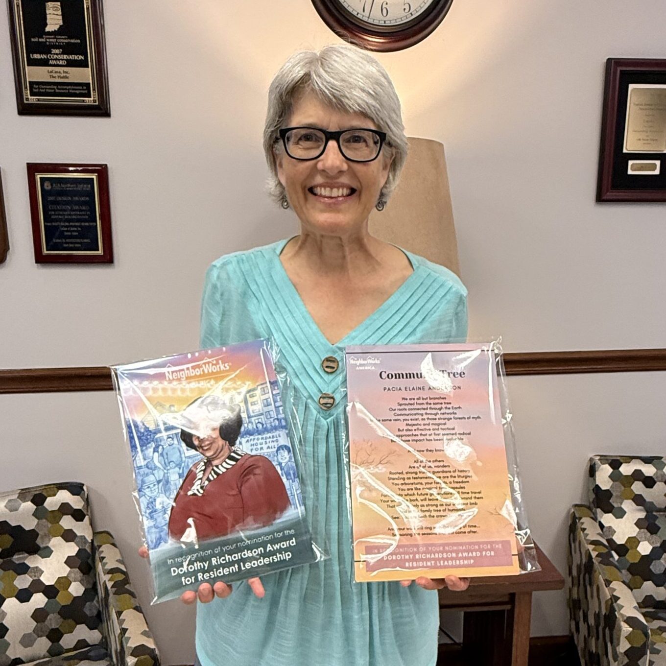 Smiling older woman in glasses and a turquoise shirt holding two colorful books in an office with plaques on the wall.