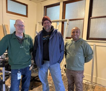 Three guys hanging out indoors, dressed casually. They’re standing in a room with large windows and equipment in the background.