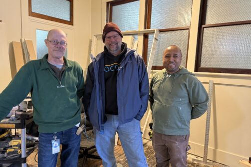 Three guys hanging out indoors, dressed casually. They’re standing in a room with large windows and equipment in the background.