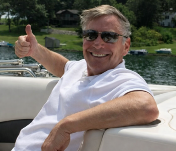Man relaxing on a boat, wearing sunglasses and a white polo, giving a thumbs up and smiling, with water and trees in the background.