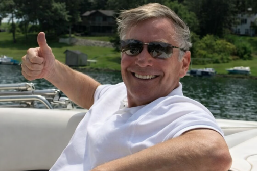 Man relaxing on a boat, wearing sunglasses and a white polo, giving a thumbs up and smiling, with water and trees in the background.