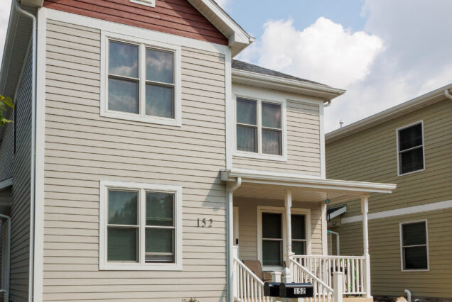 A modern beige two-story house with white trim, a small porch, and a black mailbox labeled 152. Blue sky with clouds overhead.