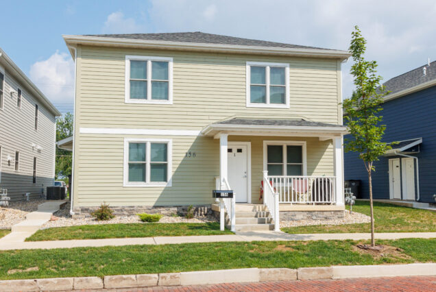 A light yellow two-story house with a front porch, a small yard, and a young tree by the sidewalk on a sunny day.