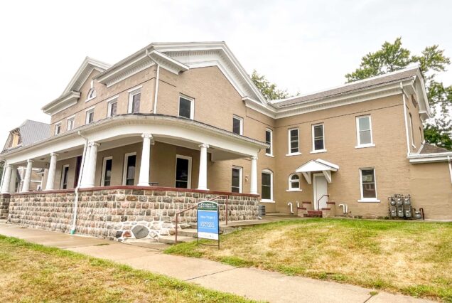 A large tan house with white trim and a stone porch, sitting on a patchy lawn under a cloudy sky.