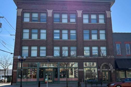 A four-story brick building with lots of windows and shops on the ground floor, sitting on a sunny street.