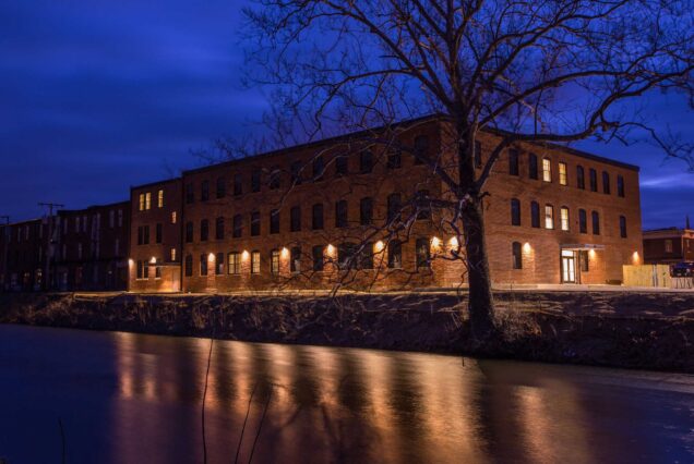 A brick building lit up at night by a river, with tree branches in front and blue evening sky above.